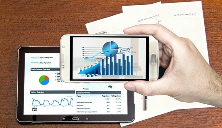 Ruka drží mobil zobrazující sloupcový a koláčový graf webové analytiky, vedle tablet s dalšími přehledy návštěvnosti.